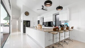 A modern kitchen with white cabinetry, a central island with bar stools, a ceiling fan, and pendant lighting. - Belle Escapes 100 Cascade Palm Cove (Palm Cove)