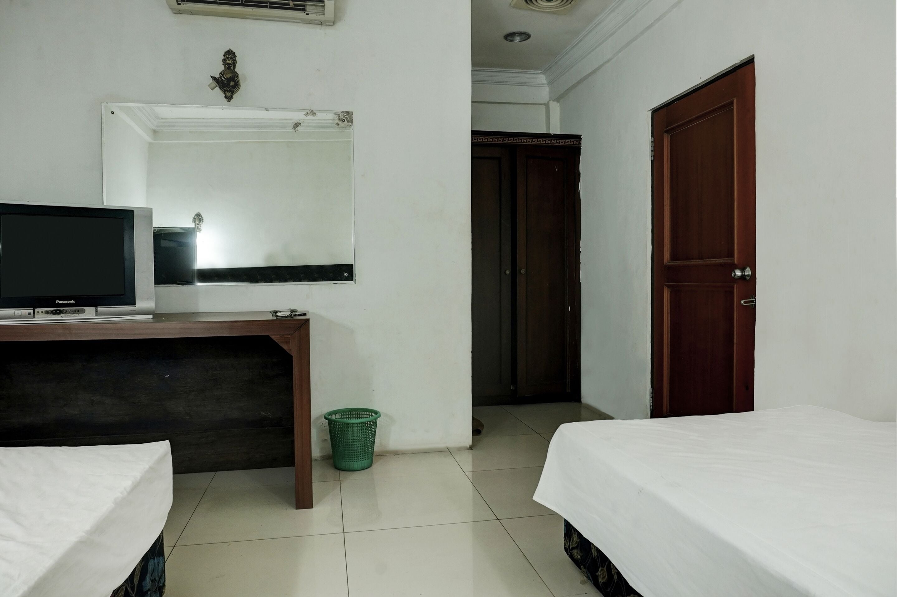 Quarto de hotel com uma cama, uma escrivaninha com televisão, um espelho, uma porta de madeira e uma lixeira verde.