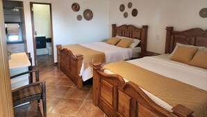 Une chambre d’hôtel avec deux lits en bois, un petit coin salon avec un fauteuil et une salle de bain visible à travers une porte ouverte.
