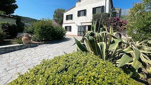 A two-story building with a stone-paved courtyard, surrounded by various plants and shrubs. - Villa Presaccia, ground floor apartment (Portoferraio)