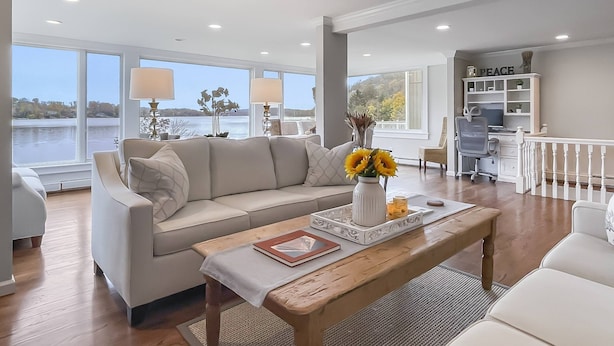 A spacious living room with a large window overlooking a body of water, a beige sofa, a wooden coffee table, and a white bookshelf with a 'PEACE' sign.