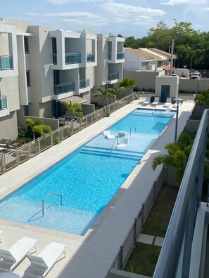 A rectangular swimming pool with clear blue water, surrounded by a white pool deck. There are lounge chairs and a small deck area with a railing. The pool is adjacent to a building with balconies and a flat roof. - Beautiful 3bed/2bath with King Beds, 2 Pools, Gym & Full Generator, near beach! (Carolina)