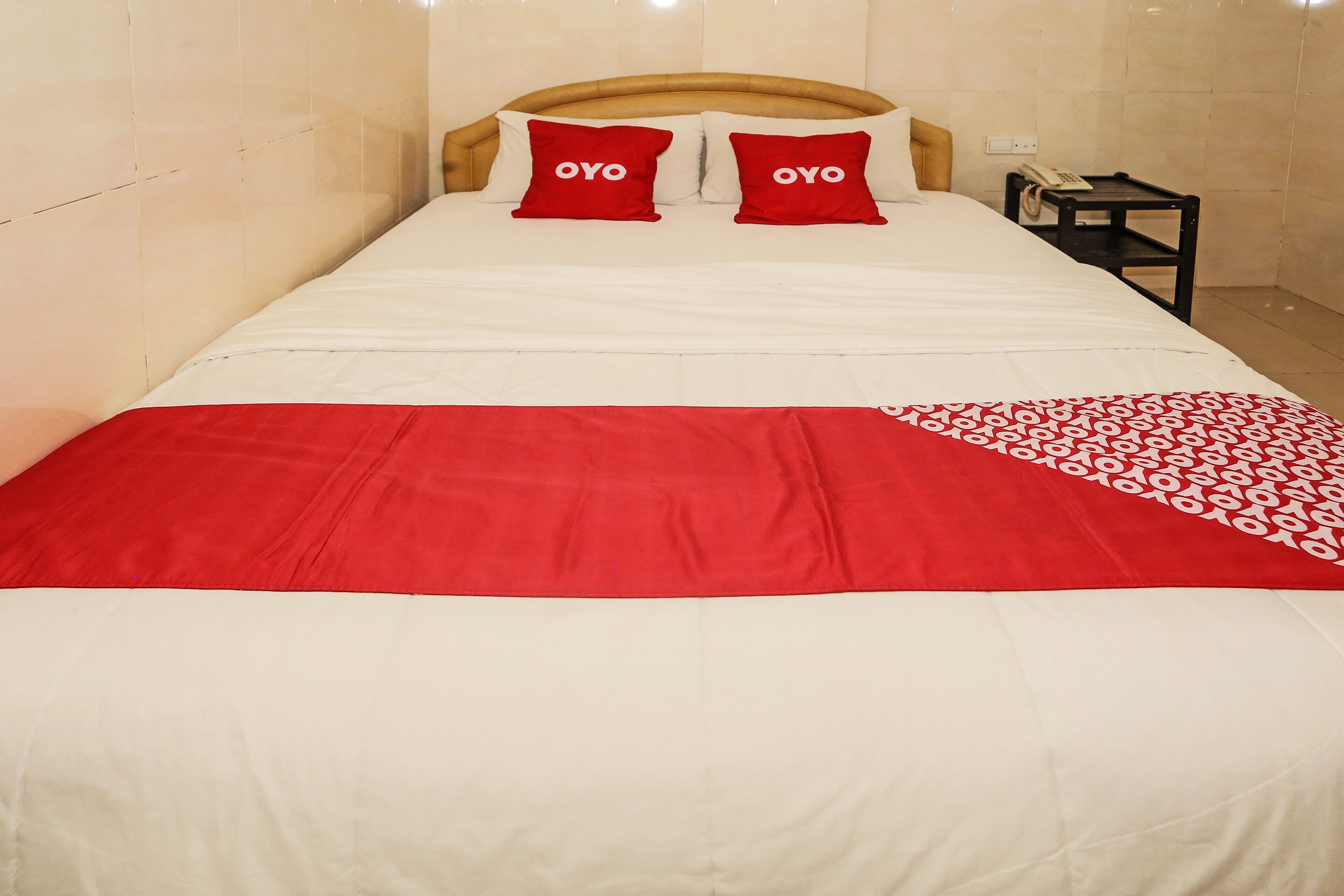 A bed with a white and red bedspread, two red pillows with the word "OYO" on them, and a small bedside table with a telephone.