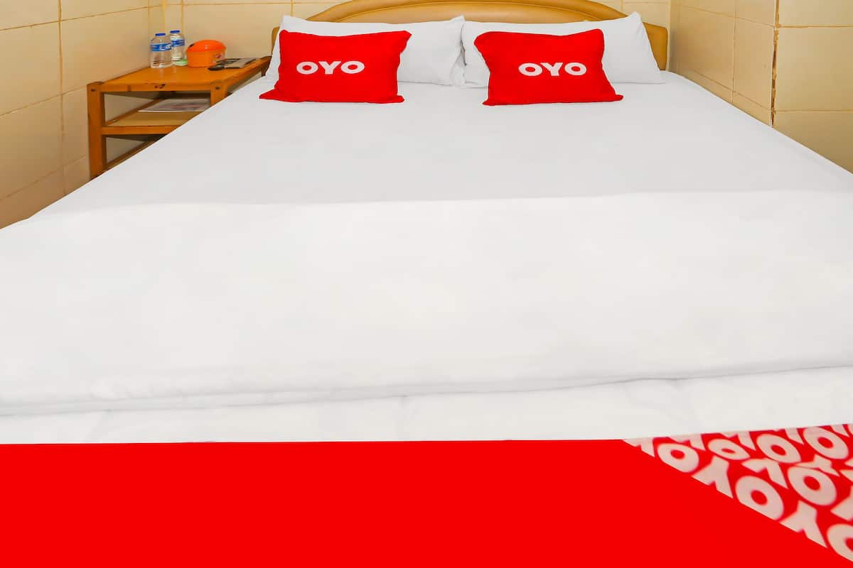 Una cama con ropa de cama blanca y dos almohadas rojas con el texto "OYO", una mesita de noche de madera con botellas y un cabecero amarillo.