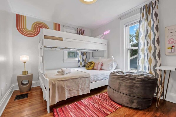 A bunk bed room with a bedside table, a lamp, a bean bag, and a window with curtains.