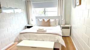 Un dormitorio ordenado con cama, mesitas de noche, un banco y una ventana con cortinas.