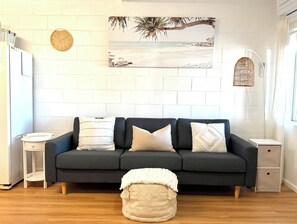 Un soggiorno moderno con un divano scuro, cuscini bianchi, un poggiapiedi rotondo, una sveglia a parete e un quadro a tema spiaggia.