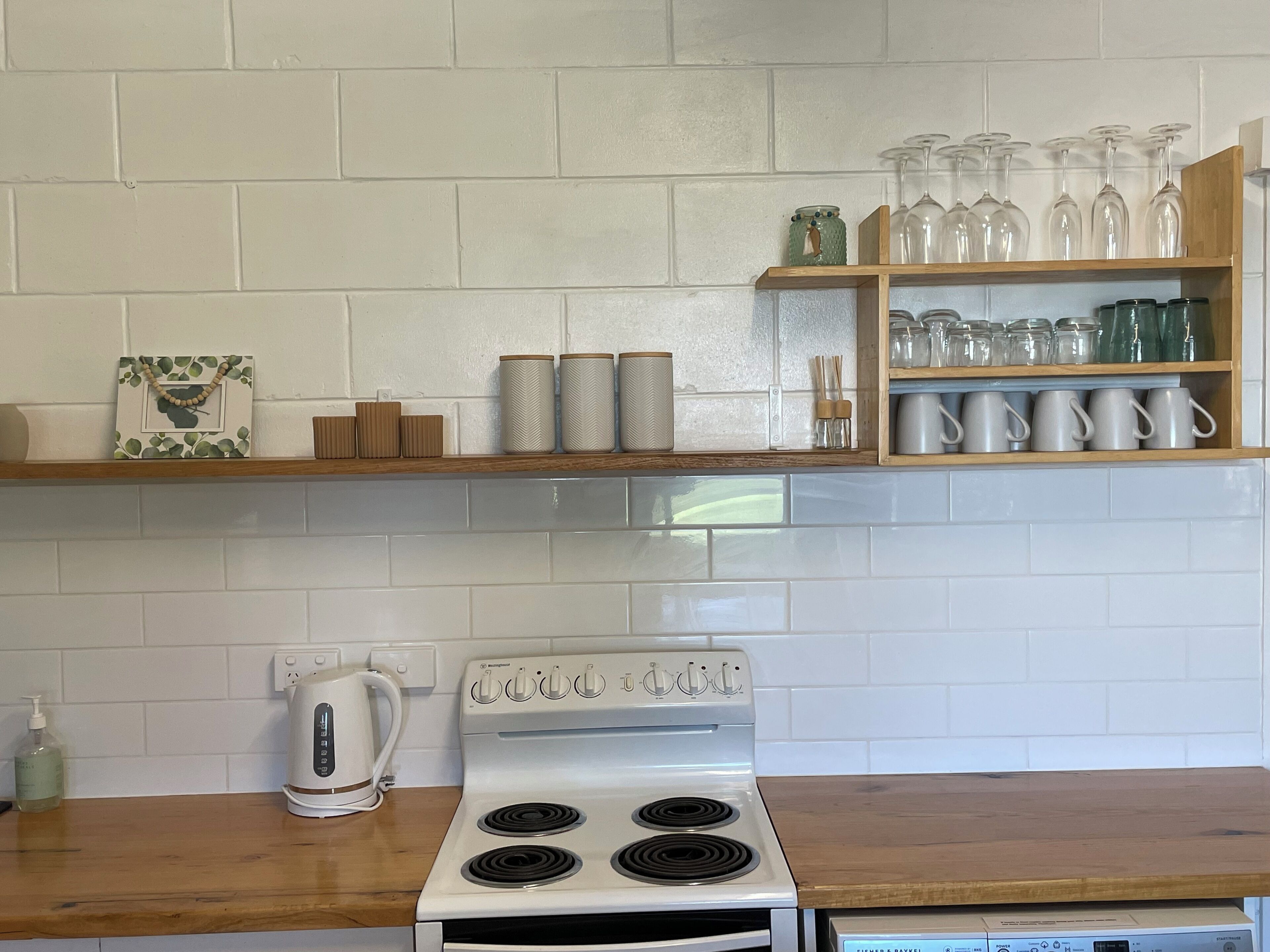 Una cocina con estufa blanca, repisas de madera con cristalería y un hervidor.