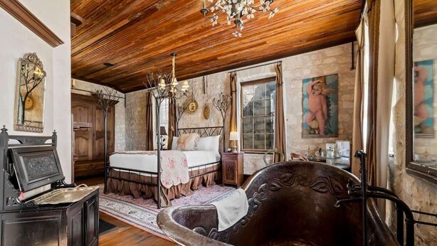 A rustic room with a large bathtub, a bed with a white bedspread, a wooden ceiling, and a chandelier. - Romantic 1BR Suite on Main Street / Prime location (Fredericksburg)