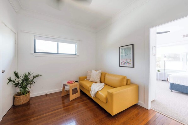 A room with a yellow sofa, a small stool, a potted plant, a framed picture, and a window.