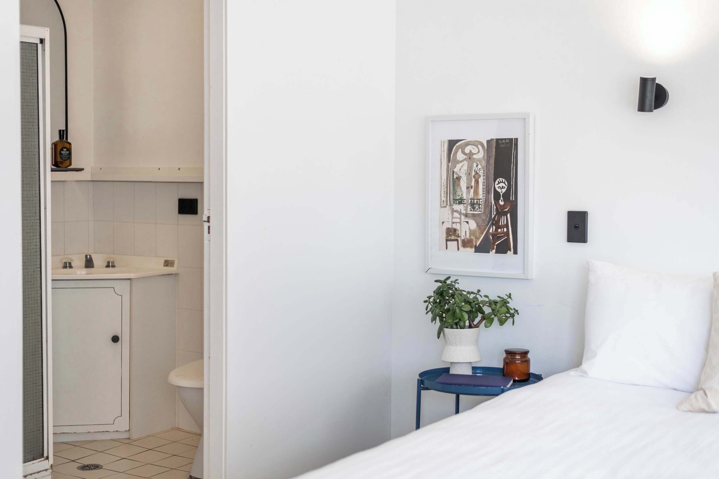 Quarto com cama, mesa de cabeceira com planta, quadro na parede e banheiro com pia e vaso sanitário visíveis através de uma porta aberta.