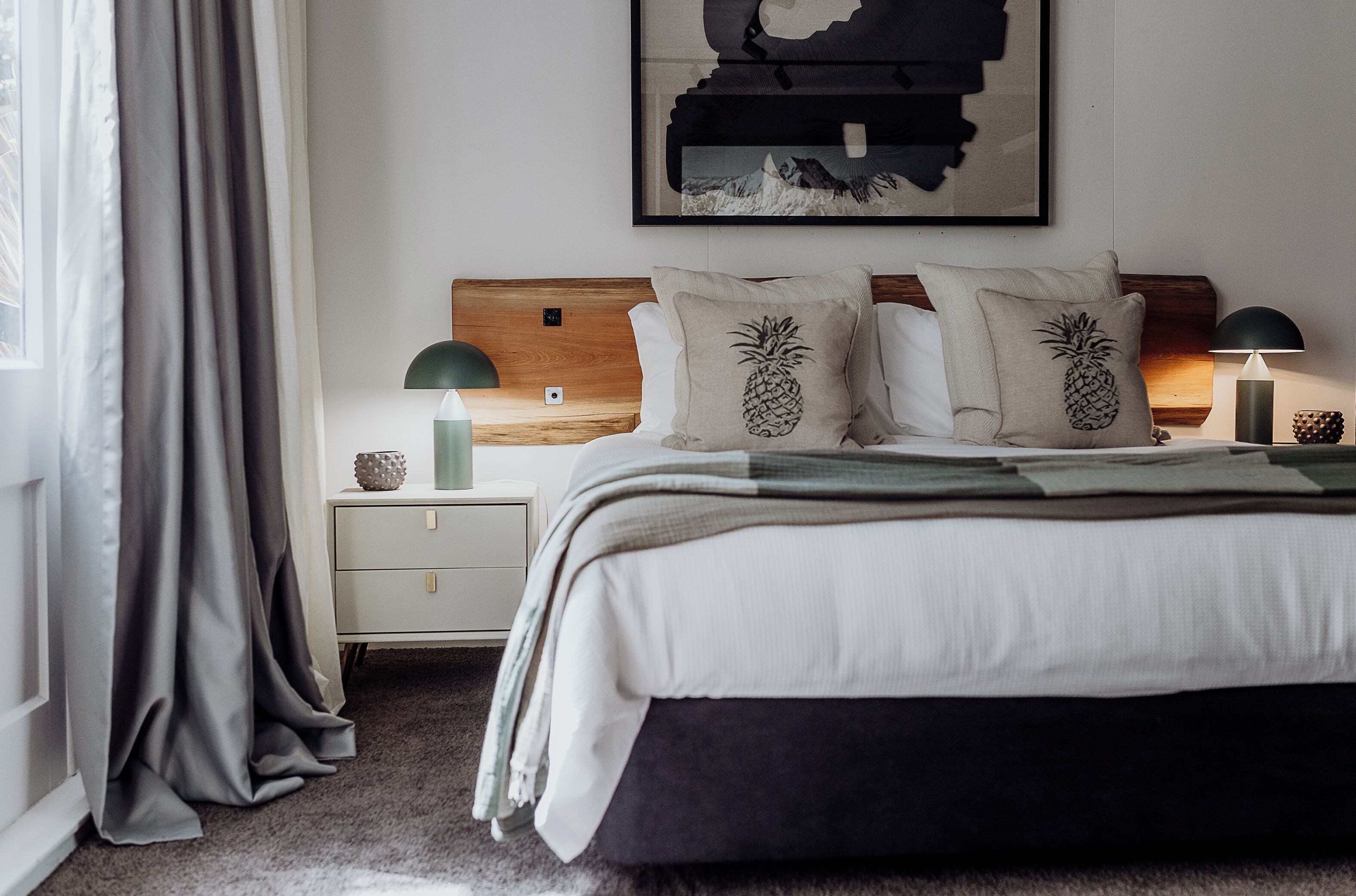 A neatly made bed with two pineapple pillows, a wooden headboard, a nightstand with a lamp, and a framed picture on the wall.