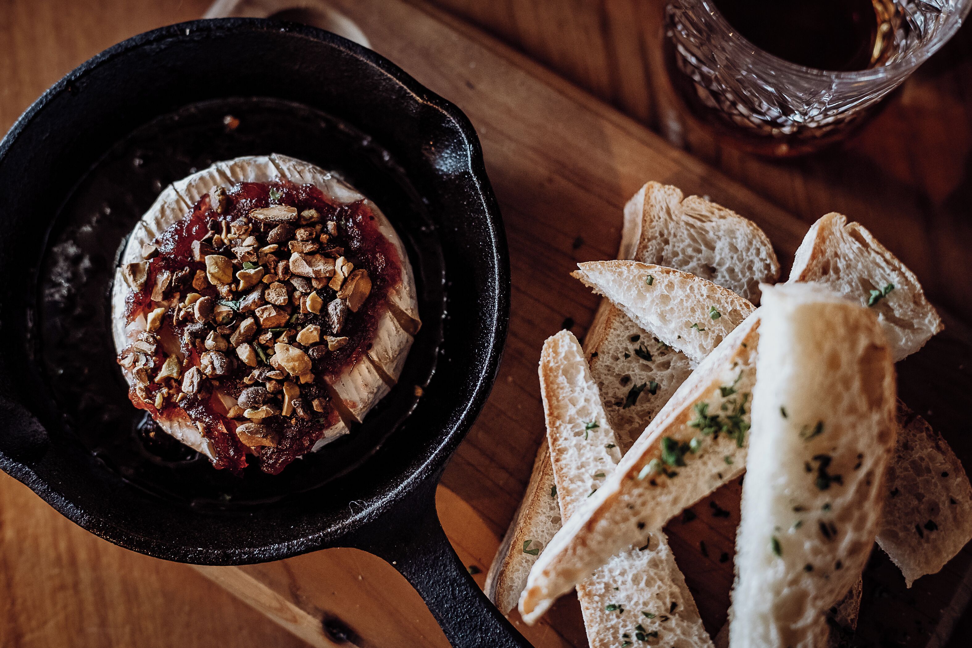 A charcuterie board with a bowl of cheese topped with nuts and a red sauce, accompanied by slices of bread on a wooden table.
