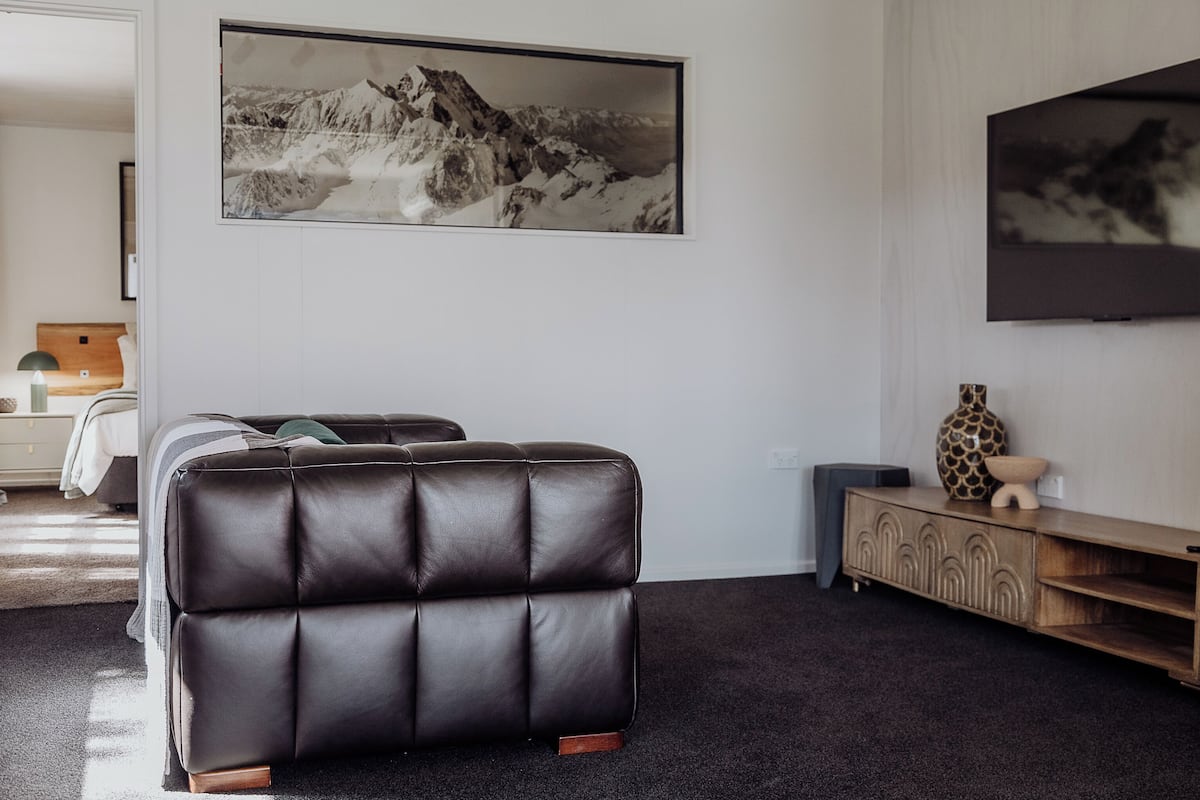 A modern living room with a leather sofa, a large framed mountain landscape, a television, and a wooden cabinet.