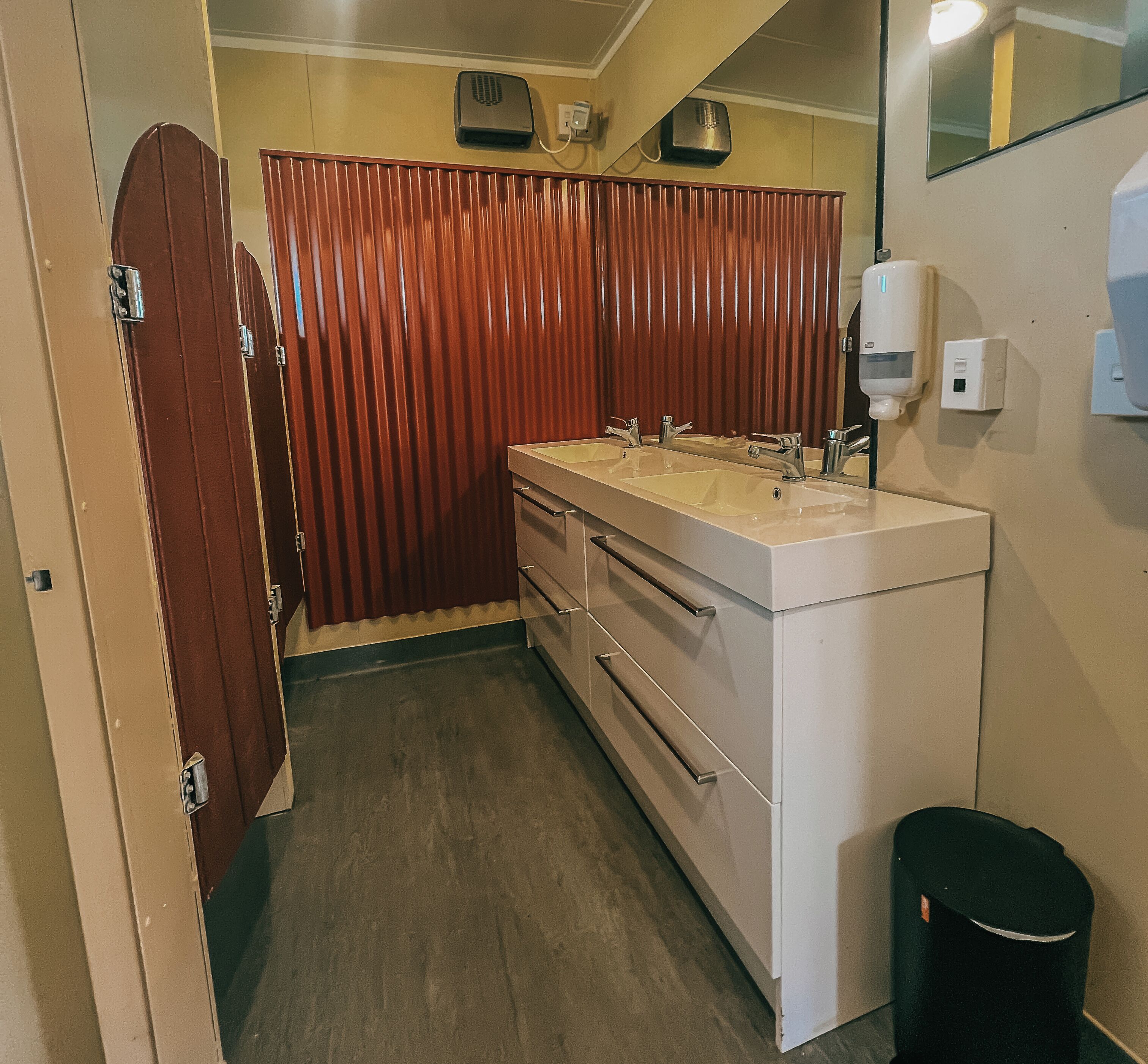 A bathroom with a double sink, a mirror, a hand dryer, and a trash bin.