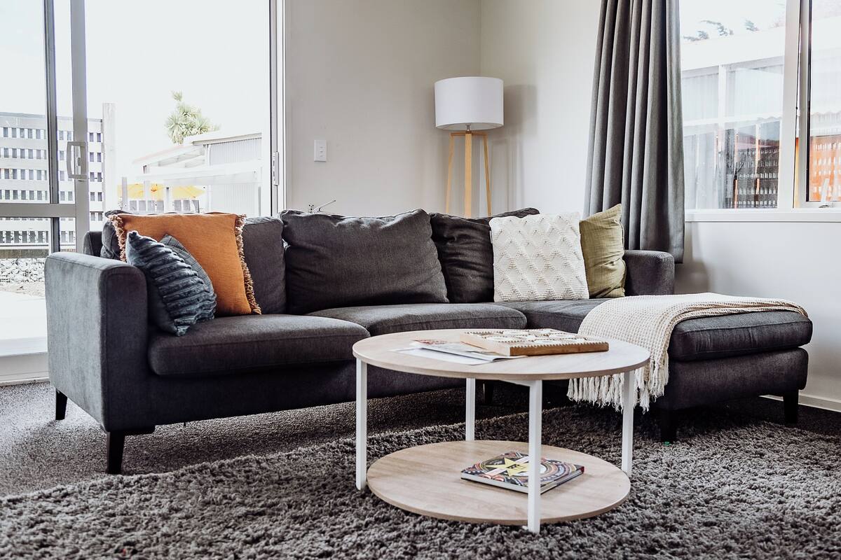 A modern living room with a dark grey sectional sofa, a round coffee table, and a large window with curtains.