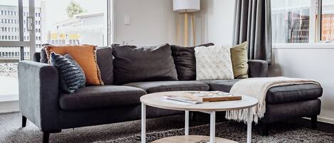 Un salon moderne avec un canapé d’angle gris foncé, une table basse ronde et une grande fenêtre avec des rideaux.