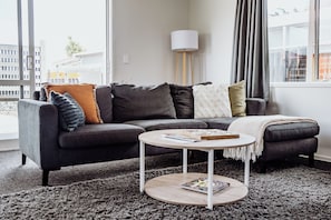 Uma sala de estar moderna com um sofá seccional cinza escuro, uma mesa de centro redonda e uma grande janela com cortinas.