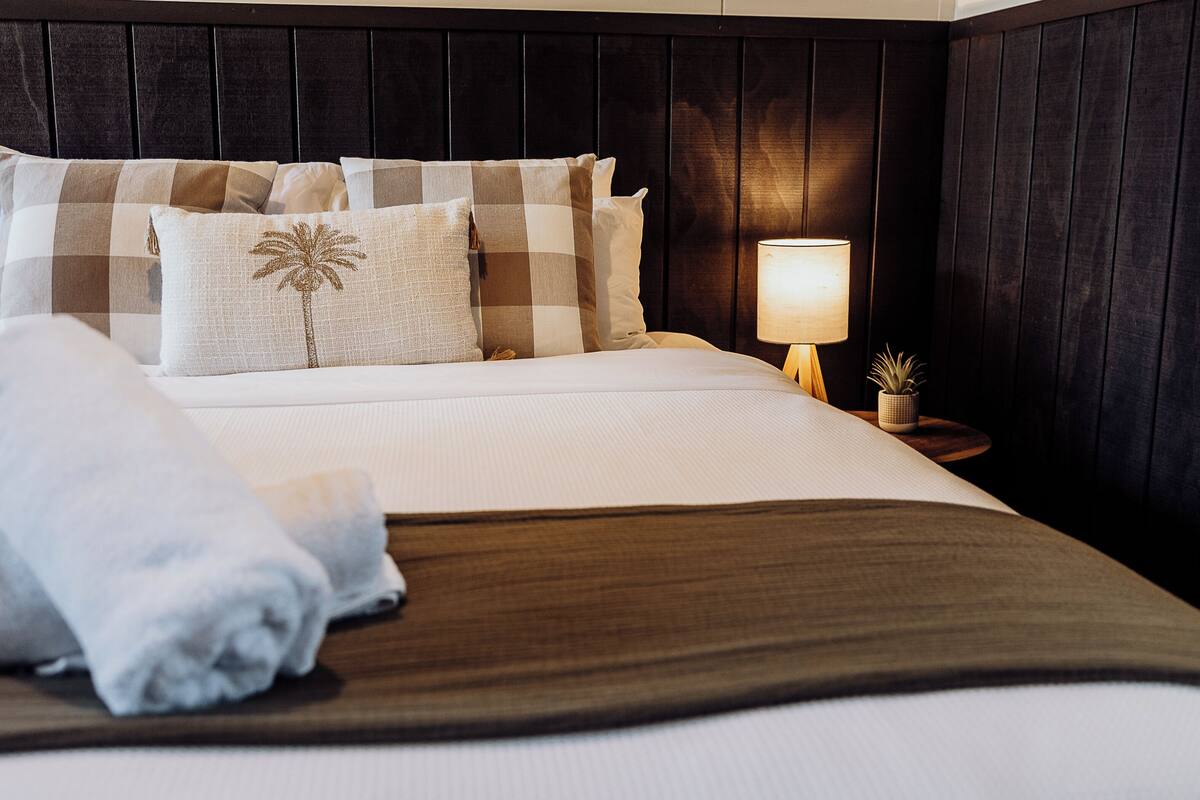 A neatly made bed with a checkered pillow set, a bedside lamp, and a small potted plant on a wooden nightstand.