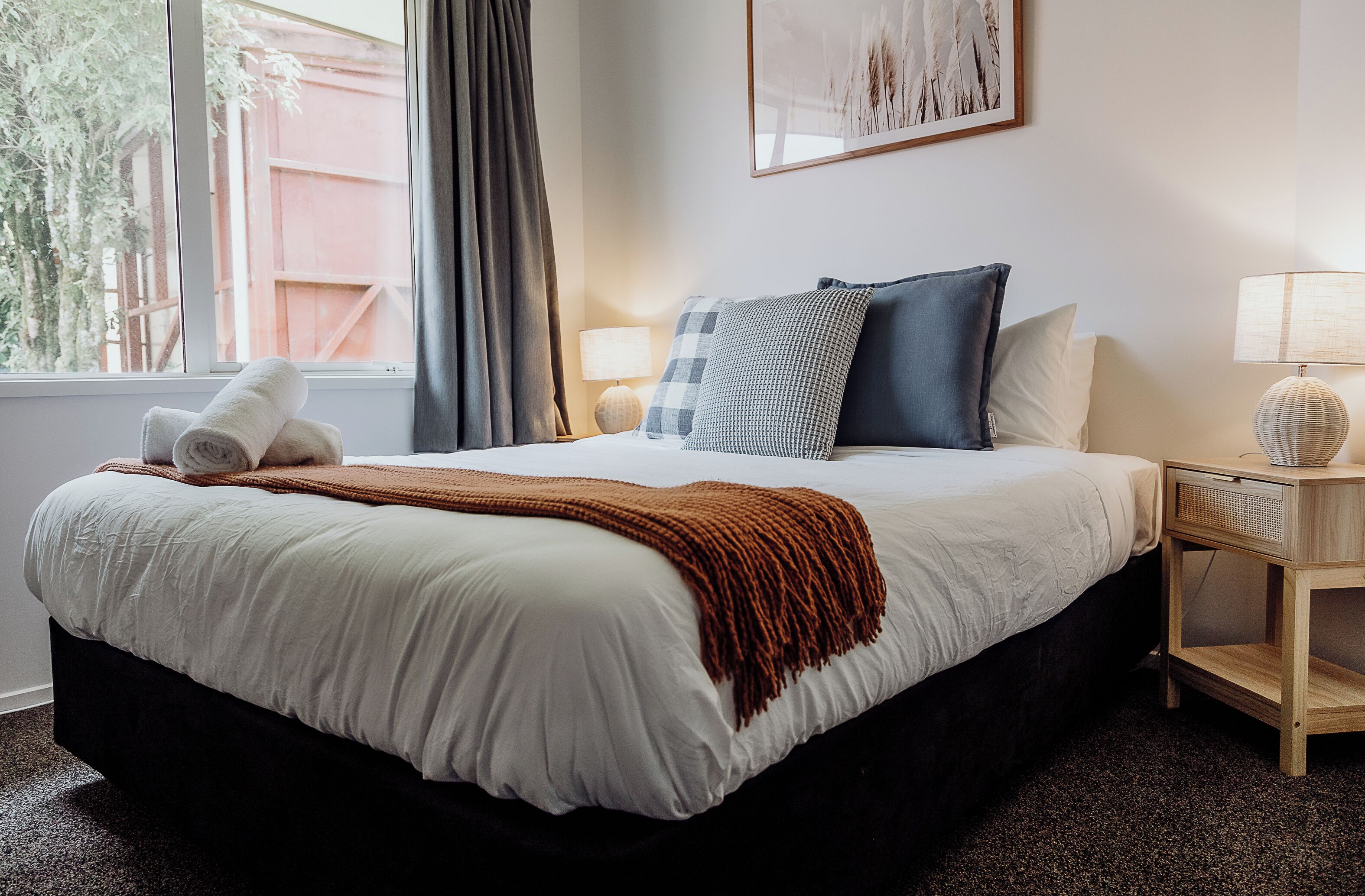 Een net opgemaakt bed met een witte dekbedovertrek, een bruin plaid en twee opgerolde handdoeken. Er staat een nachtkastje met een lamp en een decoratief object.