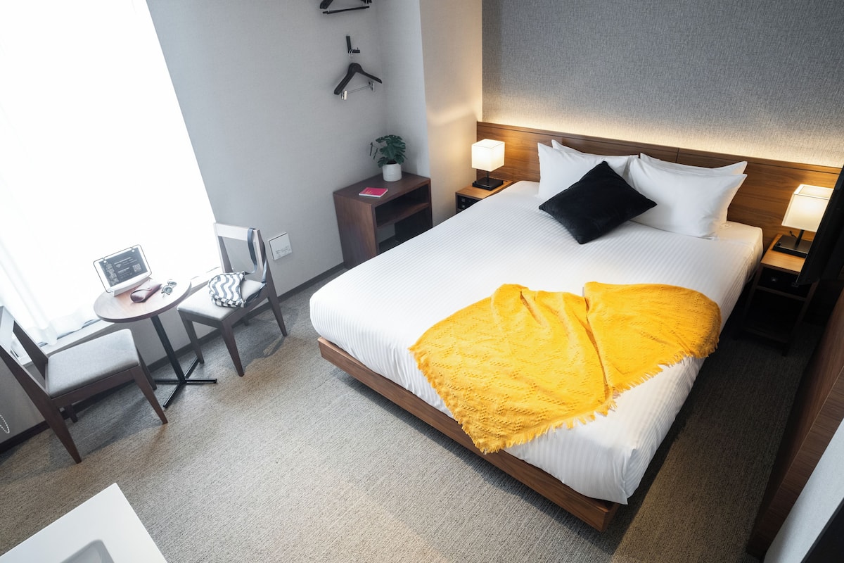 A hotel room with a large bed, a desk with a laptop, a chair, a small table, and a nightstand with a lamp.