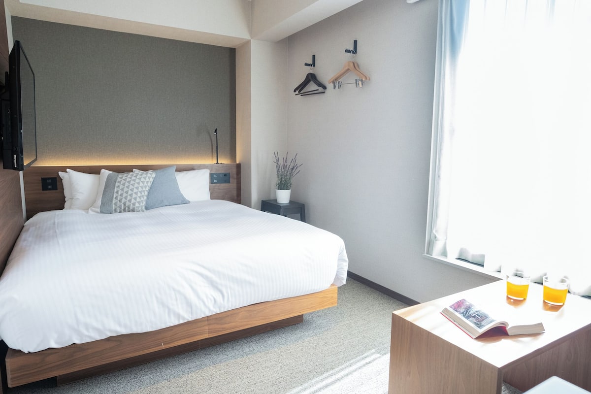 A modern hotel room with a large bed, bedside tables, a small side table with a plant, and a window with curtains.