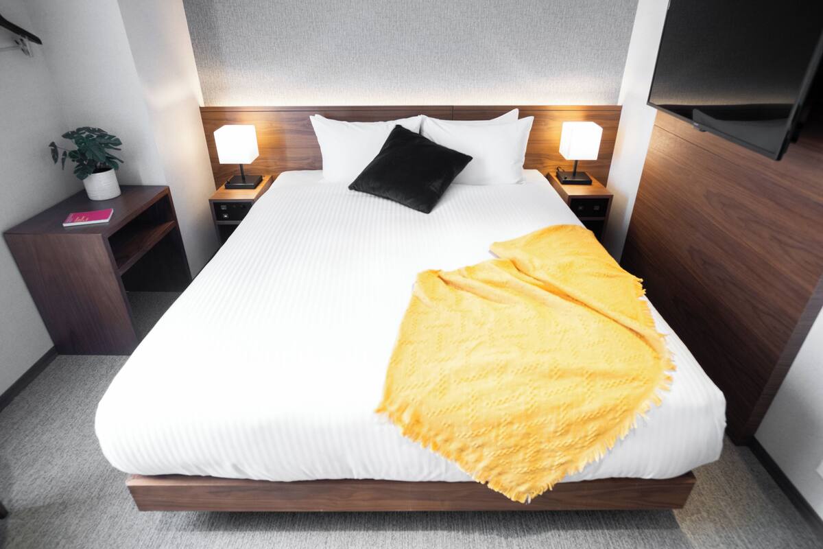 A neatly made bed with white linens, a black pillow, and a yellow throw blanket. There are two bedside tables with lamps, a small potted plant, and a flat-screen TV mounted on the wall.