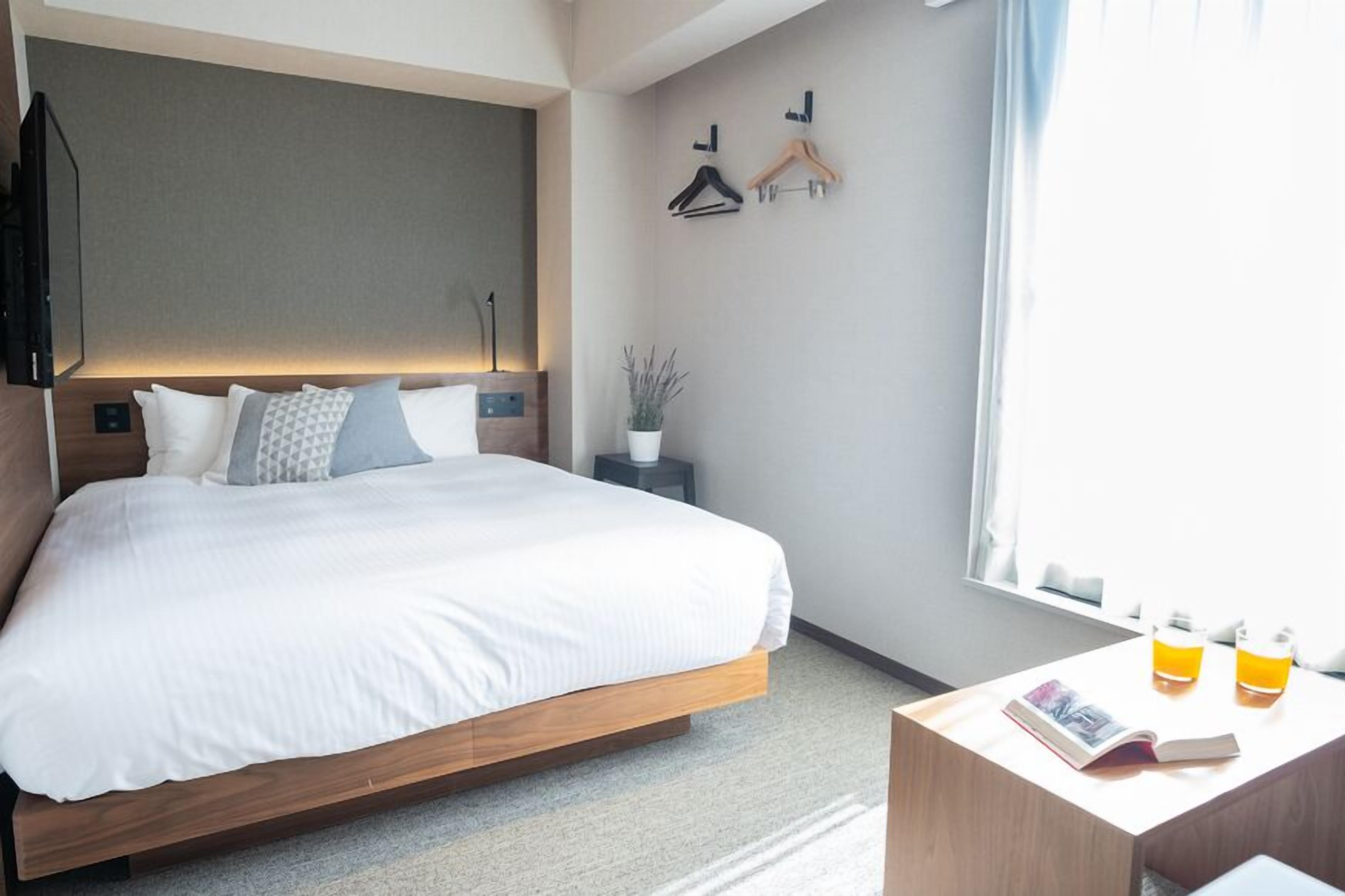 A modern hotel room with a large bed, bedside tables, a small side table with a magazine and two bottles, a potted plant, and a window with curtains.