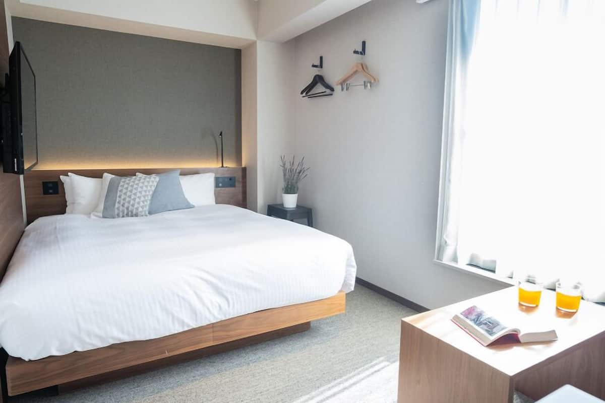 A modern hotel room with a large bed, bedside tables, a small side table with a magazine and two bottles, a potted plant, and a window with curtains.