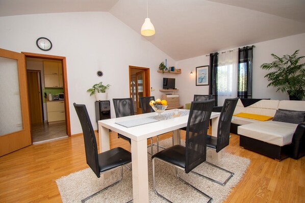 A modern living room with a dining area, a kitchen in the background, and a large window with curtains.