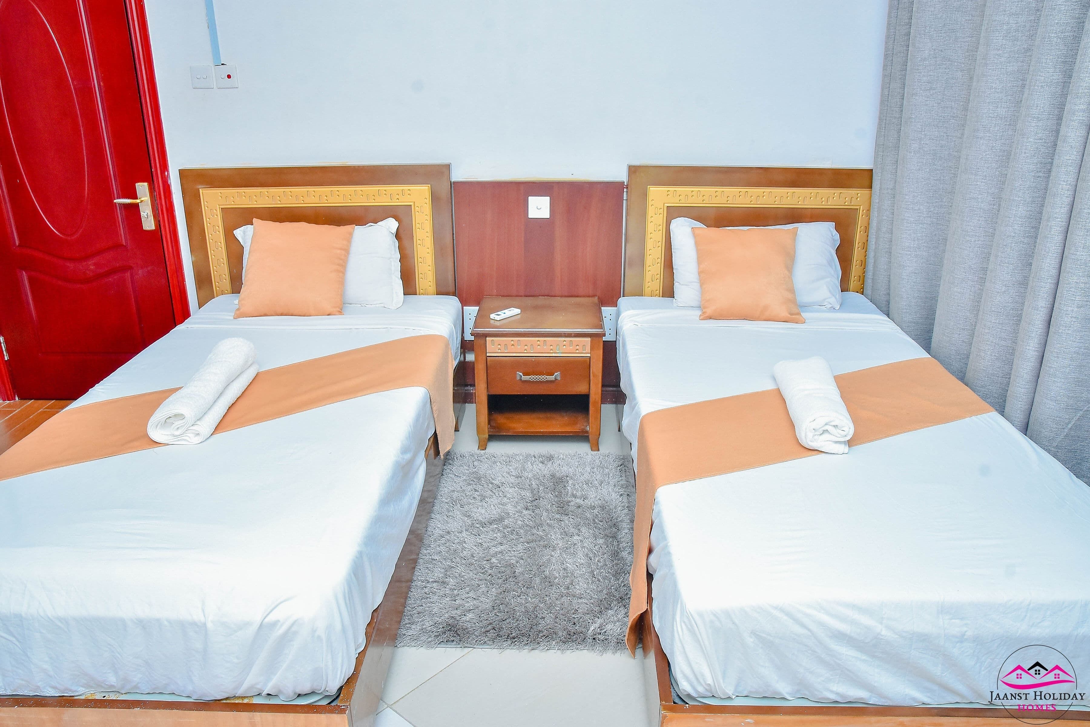 A hotel room with two single beds, a wooden nightstand, a red door, and a grey curtain.