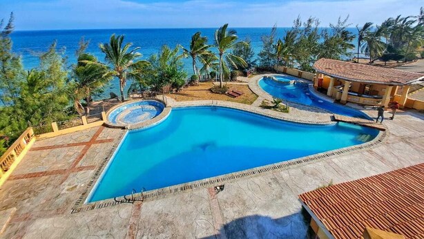 A large, clear blue pool with a curved edge, surrounded by palm trees and a view of the ocean. - Cool Breeze Beachfront Apartments (Mombasa)