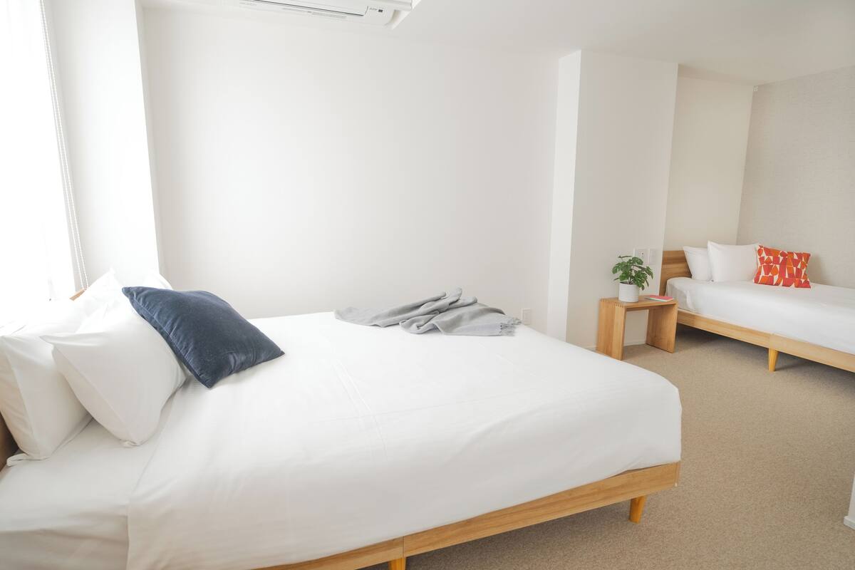 A modern bedroom with a large bed, a smaller bed, a bedside table with a lamp, and a potted plant.