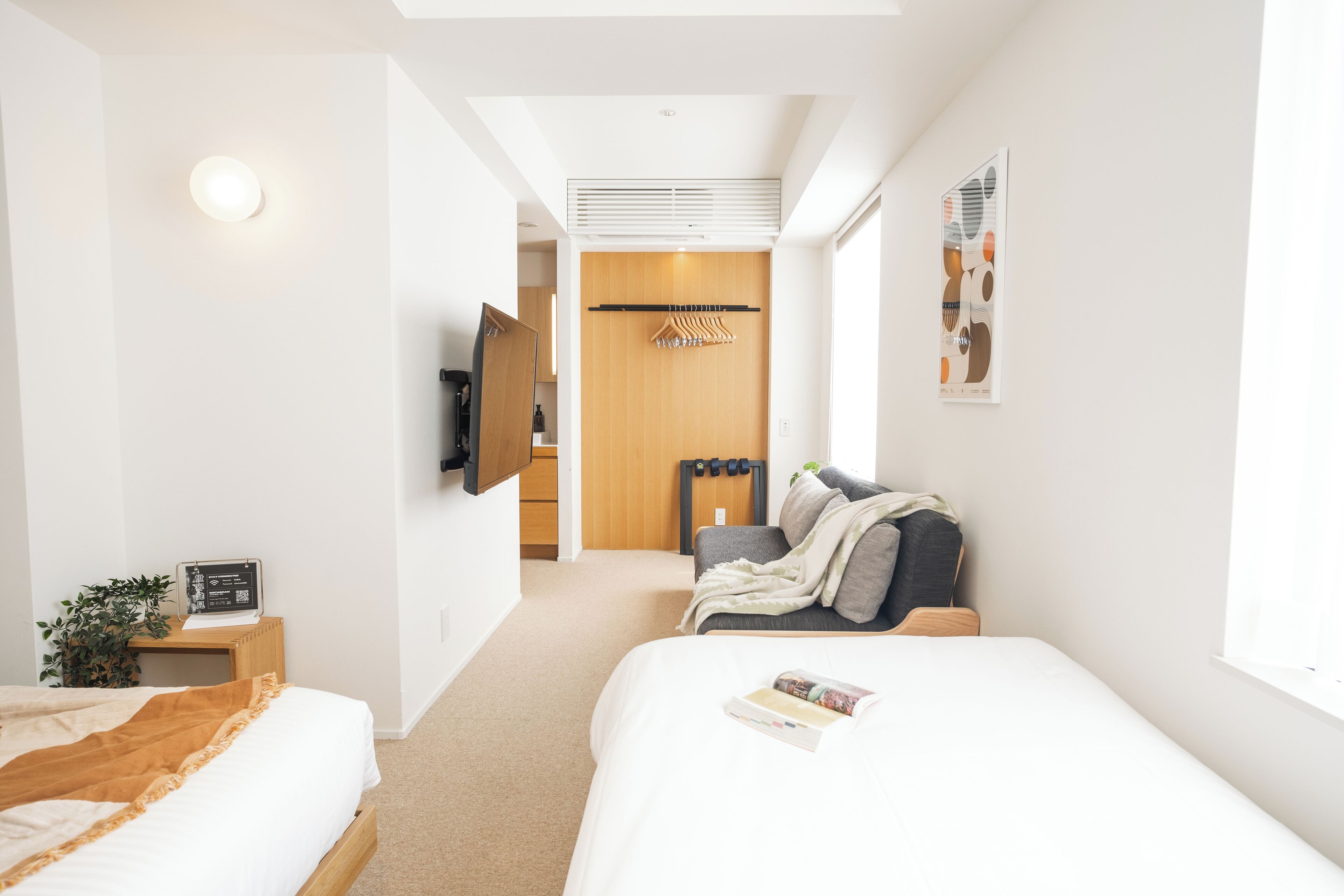 A modern hotel room with two beds, a TV, a wooden wardrobe, a coat rack, and a window with a view.