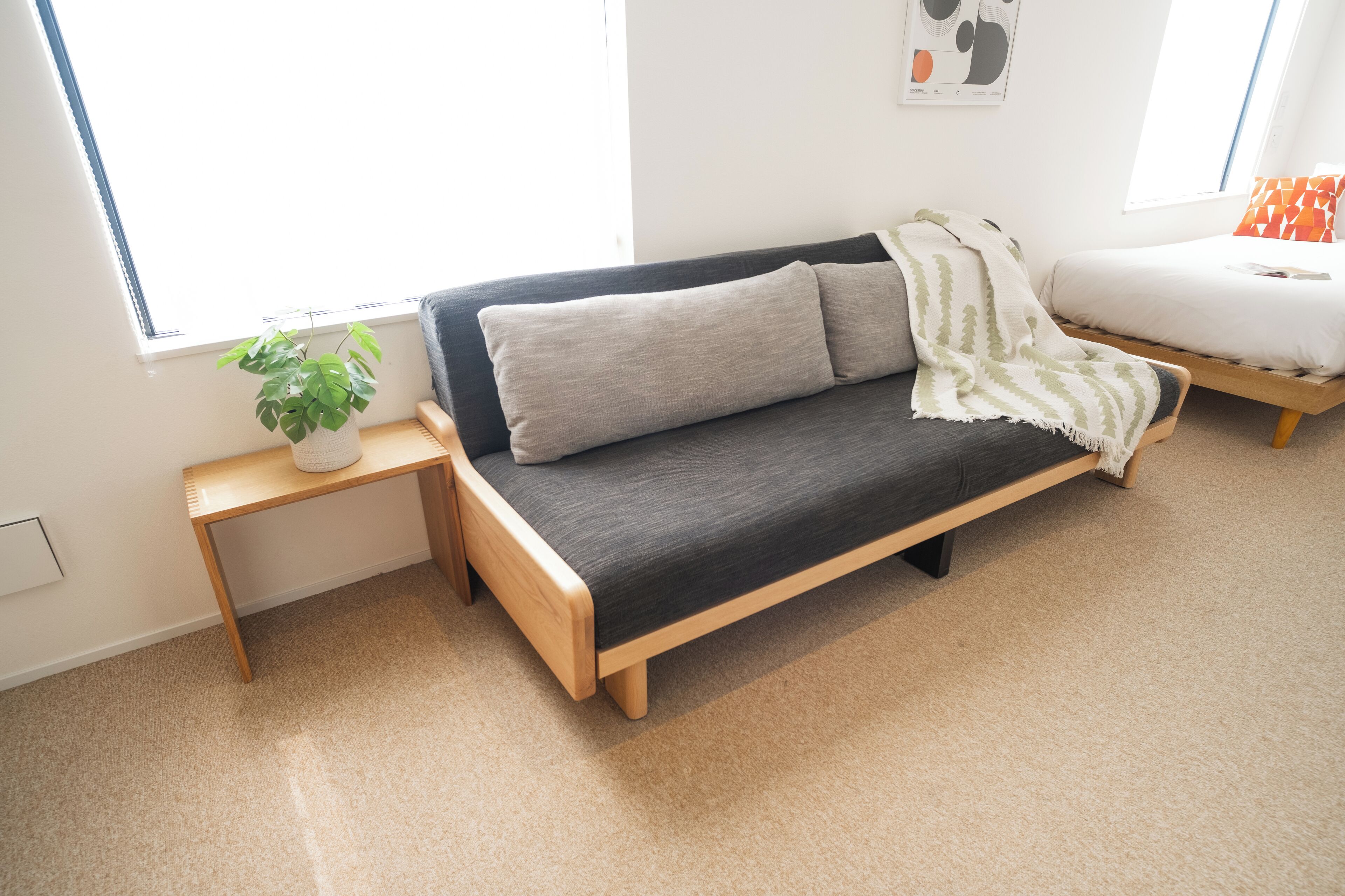 A modern room with a wooden sofa, a small side table with a plant, and a bed with a white mattress and orange pillow.
