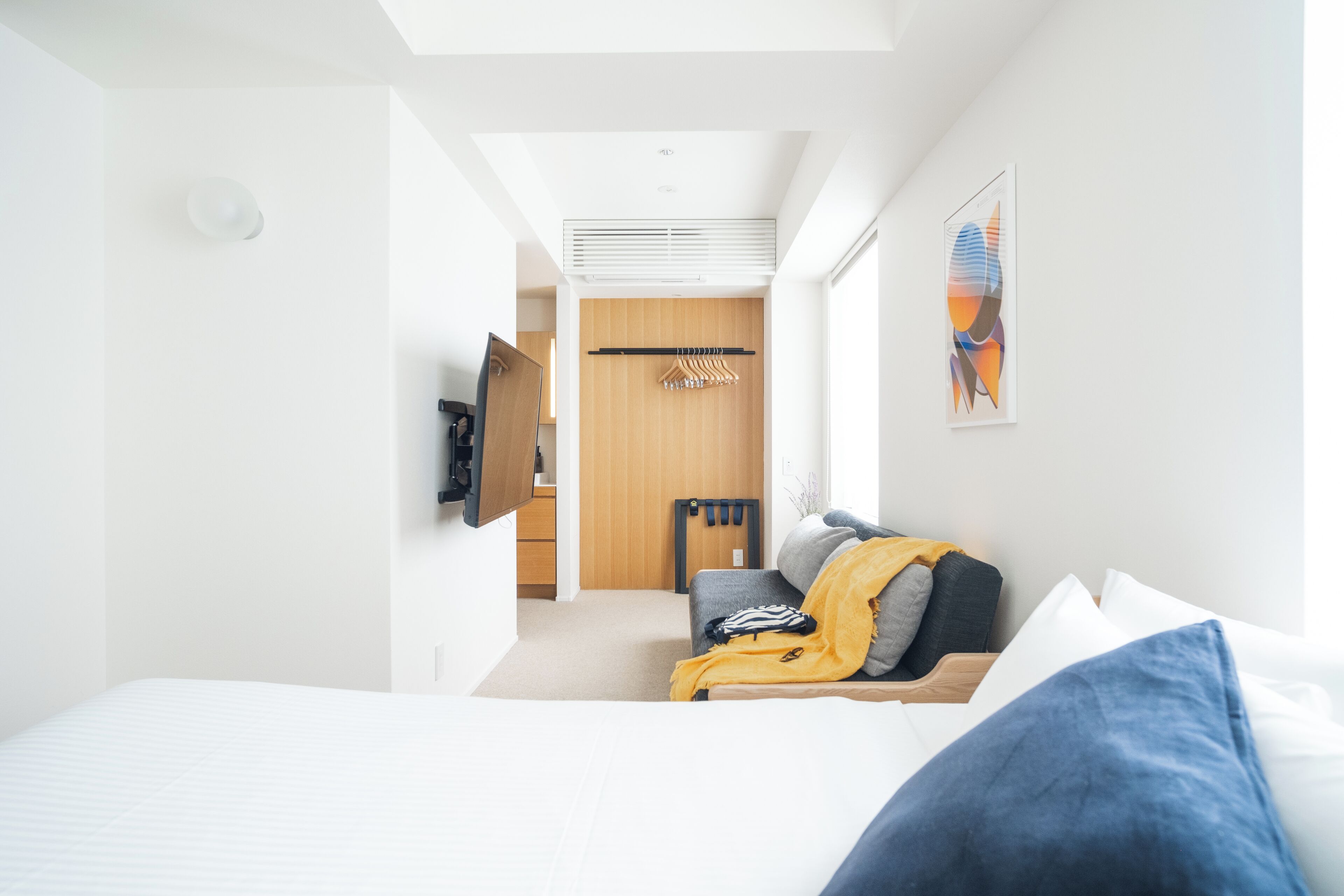 Une chambre d’hôtel moderne comprenant un lit, un coin salon avec un canapé et un fauteuil, un téléviseur fixé au mur et une porte en bois donnant sur une autre pièce.
