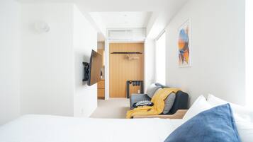A modern hotel room with a bed, a seating area with a sofa and a chair, a TV mounted on the wall, and a wooden door leading to another room.