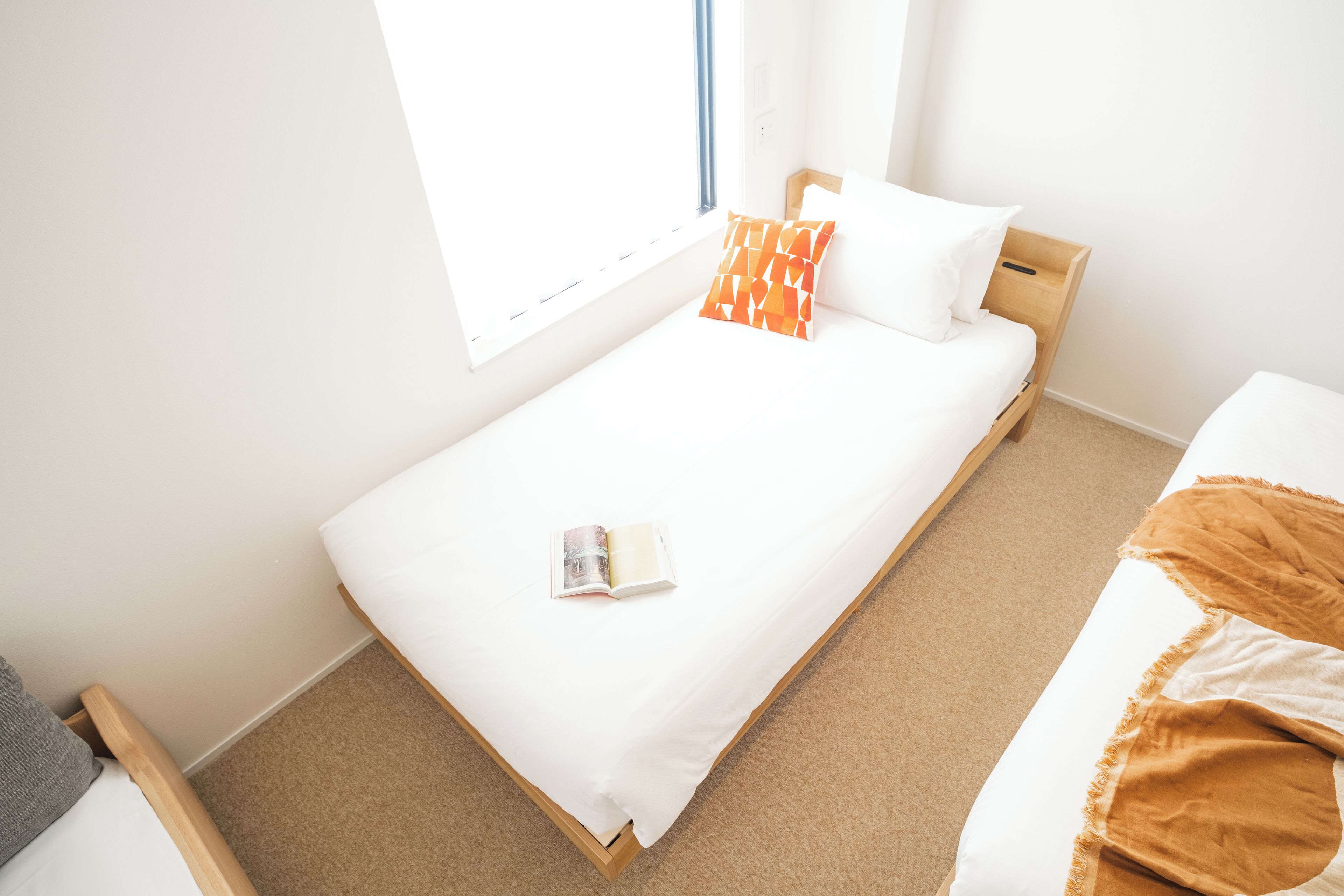 Un lit bien fait, avec un matelas blanc, un seul oreiller orange et blanc, et un livre posé sur le lit.