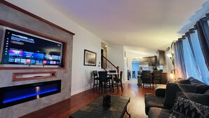 Una sala de estar con un televisor de pantalla plana montado en la pared, un comedor con sillas y una mesa, y una cocina al fondo.