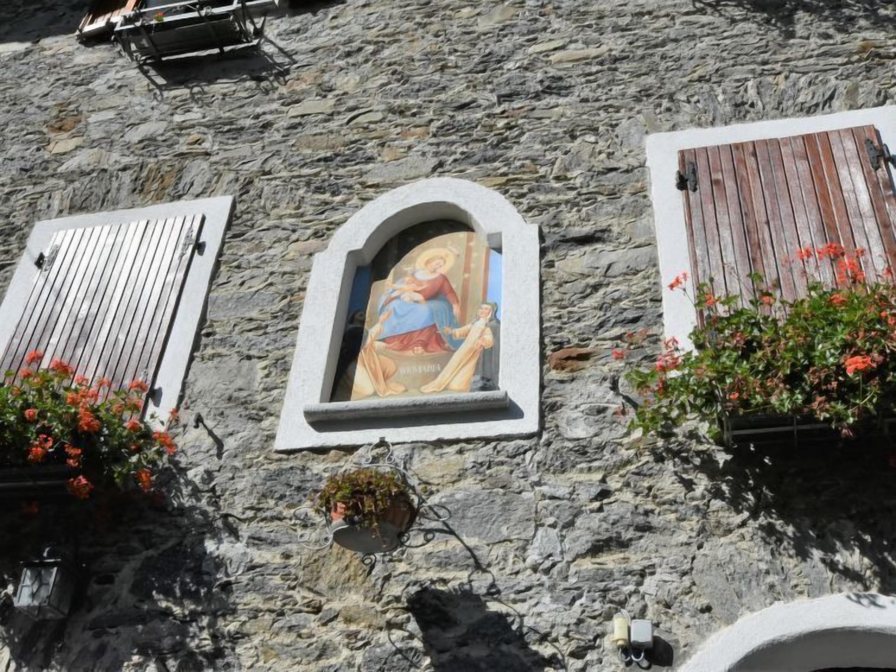 Un edifici de pedra amb una icona religiosa en una fornícula, persianes de fusta i plantes en testos.