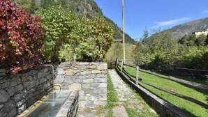 Un elemento decorativo de piedra con fuente de agua y barandilla de madera, rodeado de follaje y con montañas al fondo.