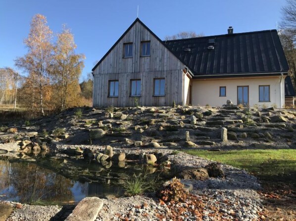Ein zweigeschossiges Holzhaus mit geneigtem Dach, gelegen auf felsigem Gelände mit einem kleinen Teich im Vordergrund.