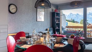 Un comedor con una mesa lista para dos personas, un reloj en la pared y vistas a montañas a través de amplios ventanales.