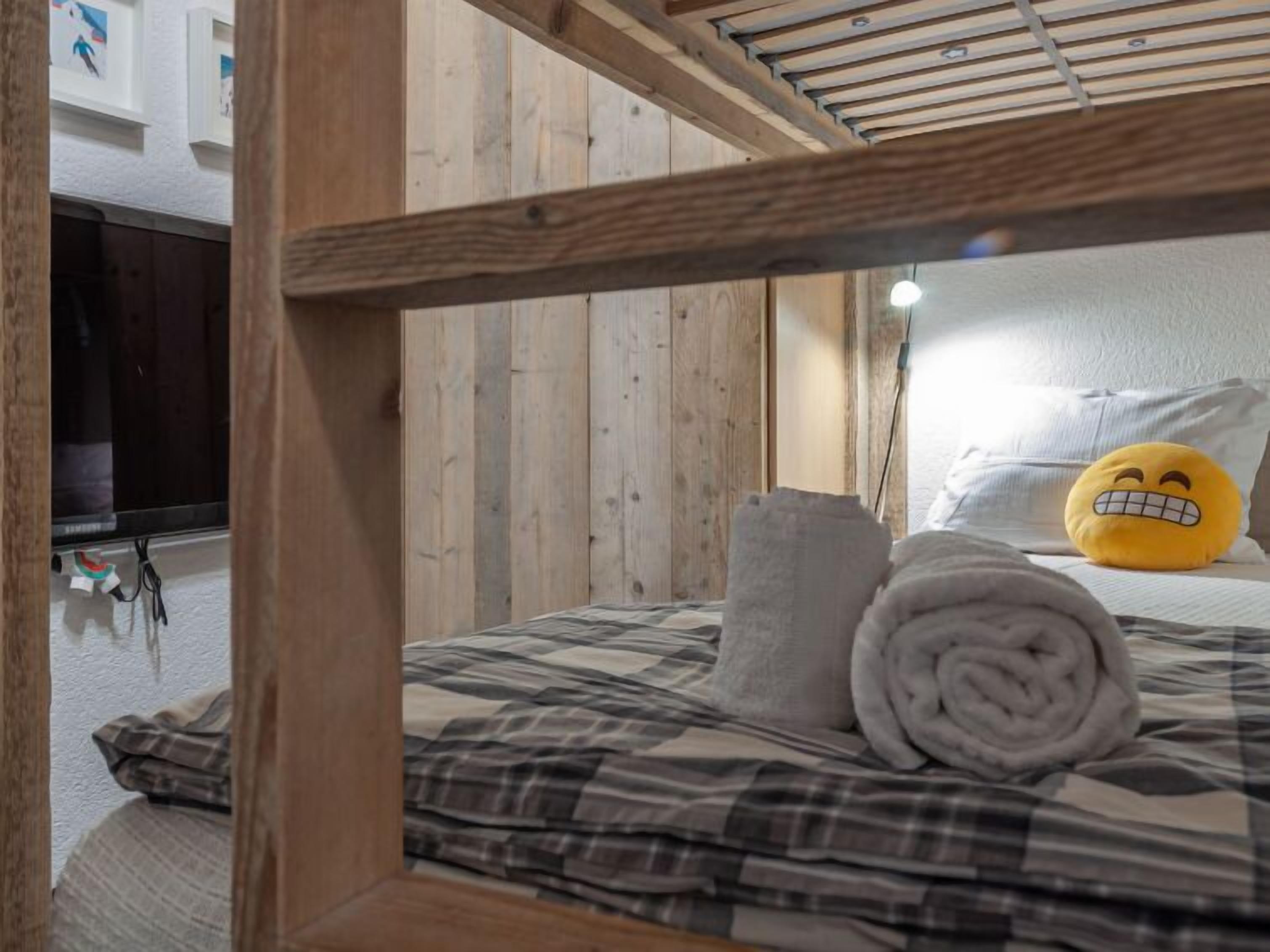 Uma cama de beliche com cabeceira de madeira, uma toalha enrolada e um travesseiro com um emoji sorridente.
