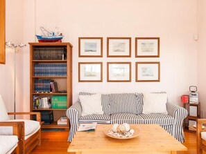 Een gezellige woonkamer met een gestreept bankstel, een houten salontafel met decoratieve objecten, een boekenplank vol boeken, en ingelijste foto’s aan de muur.