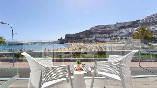 Een balkon met twee witte stoelen en een kleine tafel, met uitzicht op een strand en een kuststreeatsel.