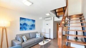 Een moderne woonkamer met een grijze bank, een salontafel en een blauw abstract schilderij. Op de achtergrond is een houten trap zichtbaar.