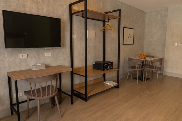 A modern room with a flat-screen TV, a wooden desk, a chair, a small table with chairs, and a shelving unit with a microwave and a hanging clothes rack.
