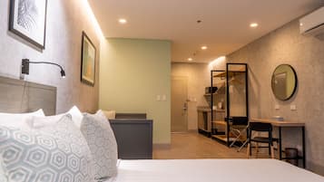 A modern hotel room with a bed, bedside table, and a desk with a chair. There is a wardrobe, a mirror, and a door leading to a bathroom.