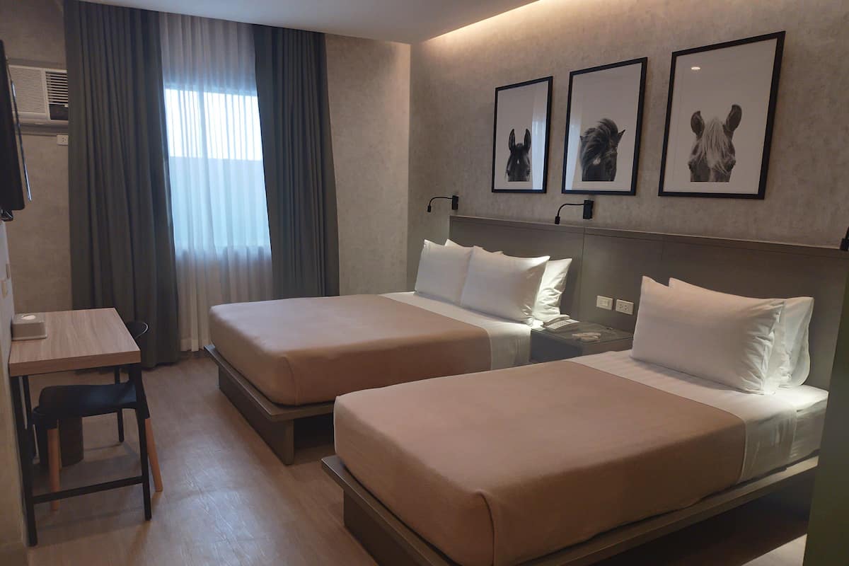 A hotel room with two beds, a desk, and a chair. There are three framed pictures on the wall.